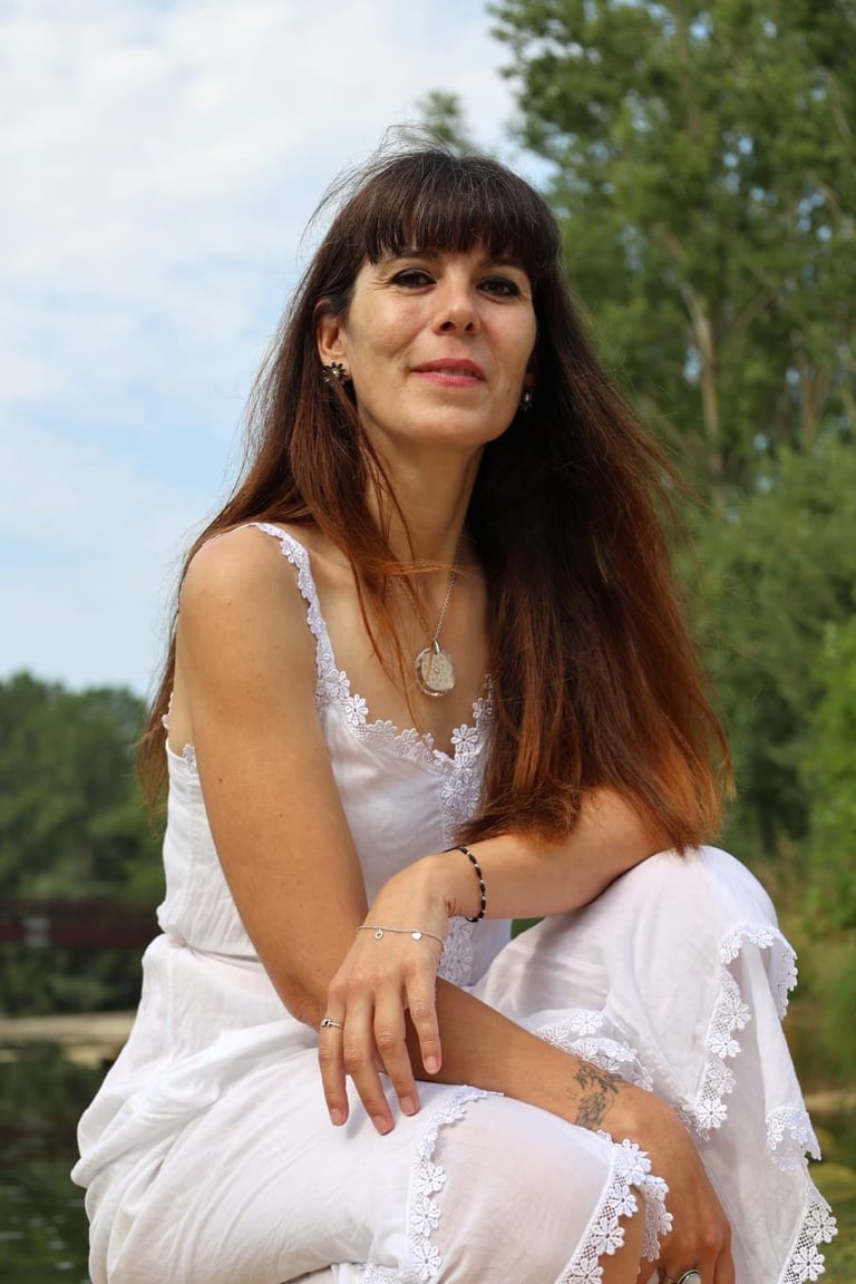 Woman with long brown hair wearing a white lace dress, posing outdoors with trees and water in background