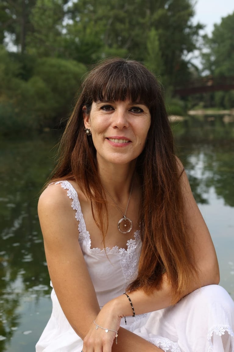 Woman with long brown hair and bangs smiling at camera by a river with trees in background