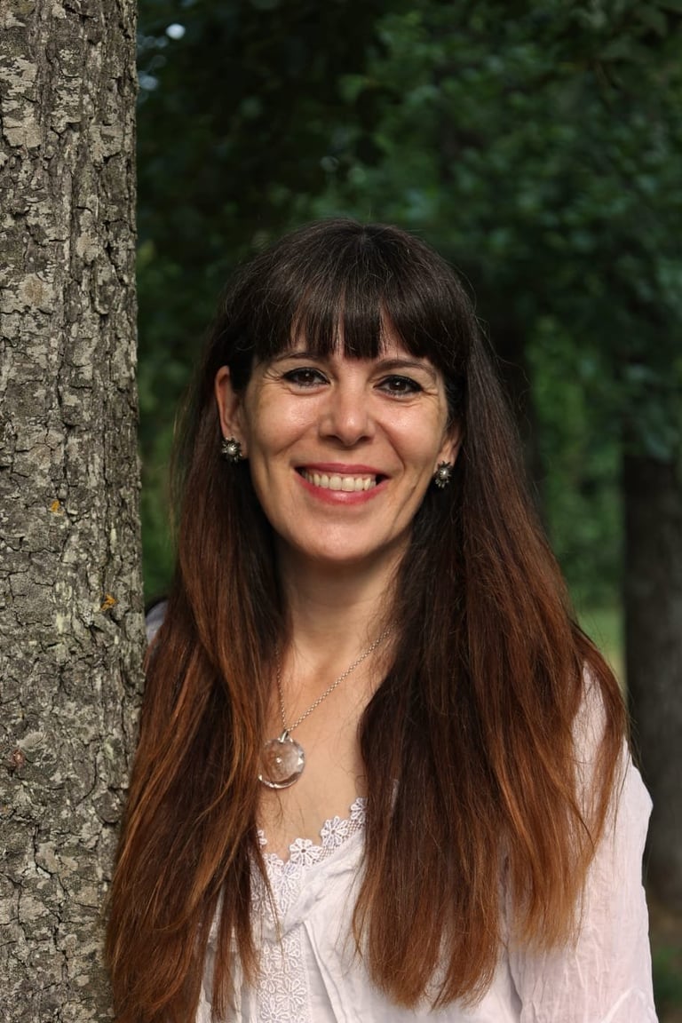 Woman with long brown hair and bangs smiling against a tree trunk, wearing a white blouse and pendant necklace